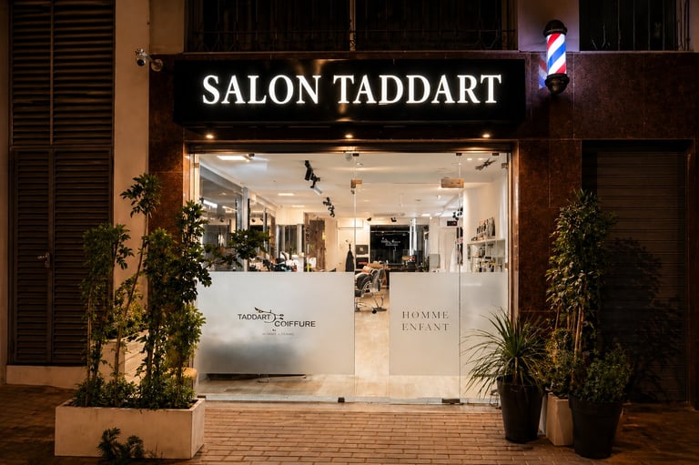 Nighttime storefront of Salon Taddart with illuminated signage, glass windows, and potted plants flanking the entrance