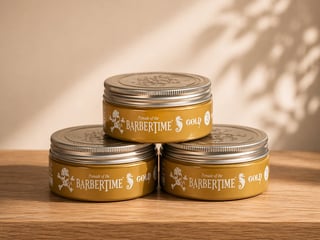 Three gold Barbertime pomade tins stacked on a wooden surface with natural lighting and window shadows in background